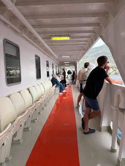 Inside a boat or ferry in the Municipal Unit of Methana, Greece, several people are present. One man wearing a black shirt and denim shorts leans against the railing. Behind him, two people also look out from the railing. Some other passengers are seated on the white seats, while others stand and walk along the red-floored passageway. Windows provide views of the outside landscape, possibly a town on a hillside.