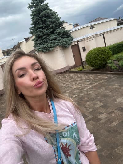 A woman with blonde hair and light skin is taking a selfie. She is wearing a pink sweatshirt with a graphic design on it and a blue strap across her chest. Her lips are pursed, and she is looking directly at the camera. The background shows a paved walkway, a tall evergreen tree, a building with a cream-colored facade, and a manicured garden. The sky is overcast. The image appears to be taken outdoors in a residential or institutional setting.