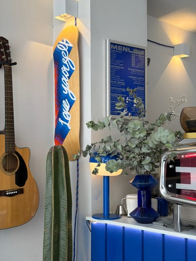 A vertically oriented indoor shot depicts a wall adorned with various items. Starting from the top, a square light fixture emits a warm glow. Below it, a blue-toned menu board lists coffee variations and prices, followed by a bouquet of eucalyptus. Adjacent to the bouquet, a vintage-style object hangs.

Further down, a skateboard with the inscription "Love yourself" is mounted above a cactus. An acoustic guitar rests below the cactus. The wall background is a plain light color. The scene takes place in San Bernardino Tlaxcalancingo, Mexico.