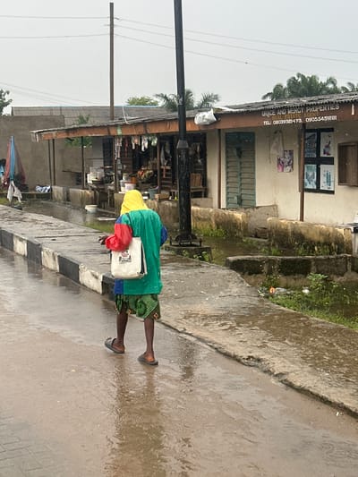 The media file depicts a person walking on a wet, paved surface, likely a street, in what appears to be a rainy or overcast day. The person is wearing a vibrant green and yellow jacket over a red shirt, and a patterned skirt. They are carrying a white bag. The setting is a roadside with a building visible to the left. The building has a corrugated metal roof and signs with text. One sign reads "GRACE AND MERCY PROPERTIES" and includes phone numbers "08039321473, 09055451281". The building's facade has windows and some posters or pictures displayed. To the right of the person, there is a grassy area with some debris and small plants. The ground is wet, with visible puddles and ripples, indicating recent or ongoing rain. The overall mood is somber due to the weather, but the bright clothing of the person adds a touch of color. The presence of overhead wires and a utility pole suggests an urban or semi-urban environment. The scene appears to be a street in Badagry, Nigeria.