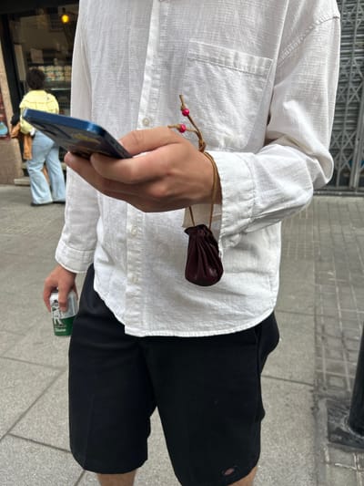 A person wearing a white linen shirt and black shorts is standing on a tiled sidewalk. They are holding a dark-colored smartphone in one hand and a green and white can in the other. A small brown leather pouch is attached to the person's wrist with a drawstring. The location appears to be urban, with a building and people visible in the background, though partially obscured by the person's hand and phone.