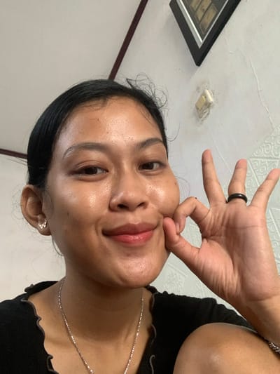 A woman is positioned at an unusual angle, seemingly taking a selfie from below. She has a dark ring on her finger and is pinching her cheek. She's wearing a black shirt with a scalloped neckline and a delicate silver necklace. The background includes a textured white ceiling, a framed picture hanging on the wall, and a light fixture.