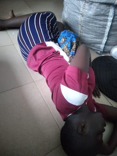 The image depicts an indoor scene in Agege, Nigeria, on a light-colored tiled floor. A dark-skinned individual lies supine with their head positioned towards the bottom right of the frame. This individual wears a maroon short-sleeved t-shirt featuring a white diagonal patch on the right shoulder/side. Their eyes are open, gazing upwards and to the right. Part of their lower body is covered by dark blue fabric with yellow and brown patterns, and one leg is bent at the knee. In the upper left corner, the bare lower leg and foot of a second dark-skinned individual are visible, alongside their upper leg covered by blue and white vertically striped trousers. A large grey woven sack rests on the floor near the upper body of the supine individual. To the right of the supine individual's head, a dark, possibly ribbed, cap or hat and a small roll of white material are positioned on the tiled surface. The floor is composed of light beige or cream square tiles with dark grout lines.