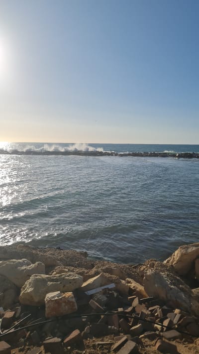 A horizontal, daytime shot of a coastline in Nahariyya, Israel. The blue sky is visible in the left of the image. Adjacent to the sky is a dark gray breakwater with small, white waves crashing against it. To the right of the breakwater is the Mediterranean Sea with visible sun glare. A rocky shoreline is to the right, with various sizes of light gray, brown, and beige rocks. There are also scattered pieces of brick, wire, and electrical cord at the edge.