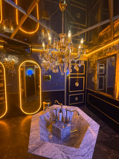 The image depicts an opulent bathroom, likely a high-end establishment, with a focus on luxurious materials and dramatic lighting. A large, ornate crystal chandelier hangs from the mirrored ceiling, casting a warm glow. The walls are a deep, dark color, possibly navy or black, with distressed textures and metallic gold accents. Recessed panels with circular gold details are visible on one wall.
Prominently featured in the foreground is a geometric sink crafted from veined marble, with a vintage-style brass faucet. On the counter of the sink, there are two bottles, likely soap and lotion dispensers. The floor is a dark, reflective material, possibly polished marble or granite, mirroring the ambient light.
To the left, an arched doorway with a vibrant orange neon outline is visible, leading into another area. Inside this doorway, a reflection in a mirror shows a dark interior with a bright blue light source. Another framed mirror with a decorative gold frame is on the right wall.
The overall ambiance is one of decadent luxury, possibly designed for a hotel, club, or a very exclusive residence. The presence of a digital display showing "55" and "22" on the ceiling suggests an air conditioning unit or similar environmental control.
The image is not from Puebla, Mexico.