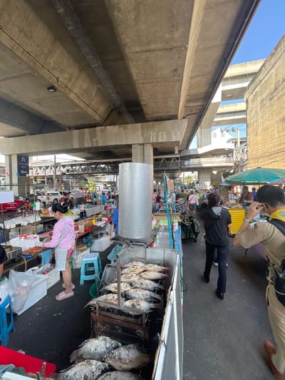 The image depicts a bustling outdoor market scene, likely in Bangkok, Thailand, under a large concrete overpass. The time of day appears to be daytime, with sunlight casting shadows.
The primary subjects are several people navigating the market. In the foreground, a woman wearing a pink shirt and patterned skirt is attending to a stall. She is wearing white gloves and appears to be handling products. Behind her, a person in a black jacket and dark pants walks away from the camera. Further back, several other individuals are visible browsing or working at stalls. One person is wearing a tan uniform with a yellow scarf, possibly a scout, and is looking at their phone.
The market is filled with various objects, most notably rows of grilled fish hanging from racks at a stall. Other stalls display a variety of goods, including food items, plastic containers, and bags. Numerous umbrellas and awnings are set up to provide shade. The setting is clearly a busy market environment, with stalls lining a walkway and a palpable sense of activity. The overpass above casts a significant shadow over parts of the market, creating a unique architectural backdrop. Electrical wires and conduits are visible running along the underside of the overpass.
The overall atmosphere suggests a lively and active market, typical of a city environment. The presence of various vendors and shoppers indicates a commercial activity is in full swing.