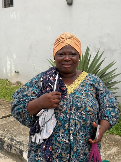 A woman smiles gently towards the camera in an outdoor setting in Igboefon, Nigeria. She is positioned against a light grey, somewhat cracked wall, with a tall, spiky-leaved tropical plant visible behind her.
The woman has a dark complexion and wears a light orange, pleated headwrap. Her attire includes a long-sleeved dress featuring an elaborate pattern of swirling motifs and paisley designs in dark blue, light blue, gold, and white on a dark background. The neckline of the dress is accentuated with a golden, embroidered trim.
In her right hand, she holds a light-colored smartphone and a purple strap, possibly from a bag. Her left hand holds a folded piece of fabric, which is dark blue with small red and white geometric patterns, resembling the fabric of another garment or accessory.
The scene is brightly lit, indicating daytime and clear weather. The woman's expression is warm and inviting. At the bottom of the frame, a concrete surface or pavement is partially visible. There is no discernible text within the image.