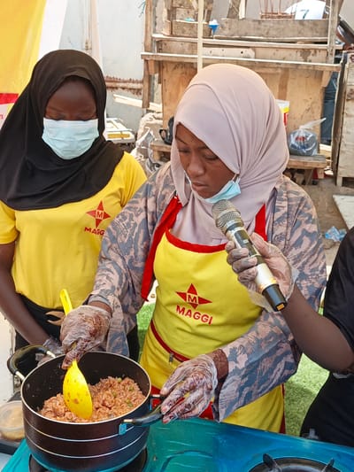 In Tarauni, Nigeria, two women are engaged in a cooking demonstration. The woman on the left wears a black hijab and a yellow t-shirt with a red "M" logo and the word "MAGGI" printed on it. She is also wearing a light blue face mask and a black skirt. The woman on the right wears a light pink hijab and a yellow apron with the same "MAGGI" branding. She is holding a microphone and wearing gloves, suggesting she is speaking to an audience or recording. Her right arm is visible, wearing a patterned sleeve. Both women are wearing transparent plastic gloves. In front of them is a black pot on a teal gas stove, containing rice mixed with peas. A yellow spatula is in the pot. The background suggests an outdoor setting with wooden structures and some debris.