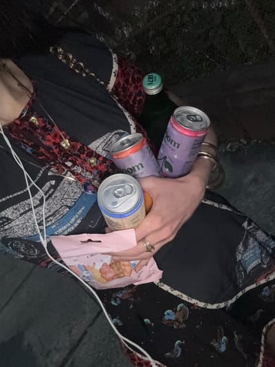 A person, seen from the chest down, sits outdoors with a dark fabric draped over their lap. They are wearing a black dress with a blue and red patterned print, along with a gold necklace. In their hands, they are holding two cans of "Bloom" energy drinks, one purple and one orange, and a small pink package of candy. A green bottle with a white cap is visible next to the cans. The person is also wearing several rings and bangles. The background consists of dark greenery and what appears to be a concrete surface. The image quality suggests it was taken at night or in a dimly lit area.