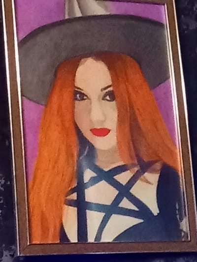 A framed, stylized portrait depicts a woman with long, bright orange hair, a wide-brimmed gray hat, and a dark garment adorned with a pentagram. Her eyes are dark, and her lips are painted red. The background is a solid purple. The image appears to be a painting or a drawing, framed in wood with a textured dark background surrounding it. The exact location in Nakhodka, Russia, is not discernible from the image itself.
