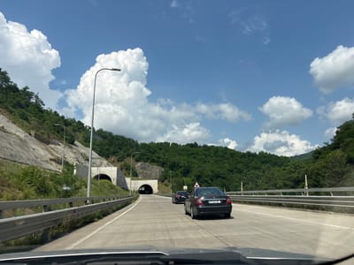 The image depicts a view from a vehicle driving on a highway. The highway curves to the right, with guardrails lining the left side and a steep, forested mountainside on the right. Ahead, the highway leads towards two tunnel entrances carved into the mountainside. The sky is a clear blue with large, puffy white clouds. Two cars are visible on the highway in front of the vehicle, with one car in the foreground and another further ahead. The scene is consistent with a highway in a mountainous region, and could plausibly be located in the Achara region of Georgia, known for its mountainous terrain and tunnels.