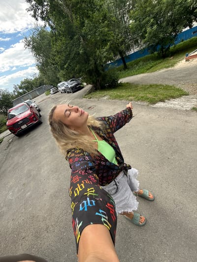 The media file shows a young woman standing on a paved road in Chelyabinsk, Russia. She is wearing a vibrant, multi-colored jacket with a repeating Chinese character pattern, a bright green tank top, and distressed denim shorts. Her feet are adorned with green slides. The woman has her eyes closed and her head tilted back, suggesting a moment of enjoyment or relaxation.
The scene appears to be outdoors, with trees and a building with blue siding visible in the background. Several cars are parked along the road, including a red SUV and darker sedans. The sky is visible with white clouds against a blue background. The lighting suggests it is daytime. The overall mood is casual and carefree. There is no other discernible text in the image besides the pattern on the woman's jacket.