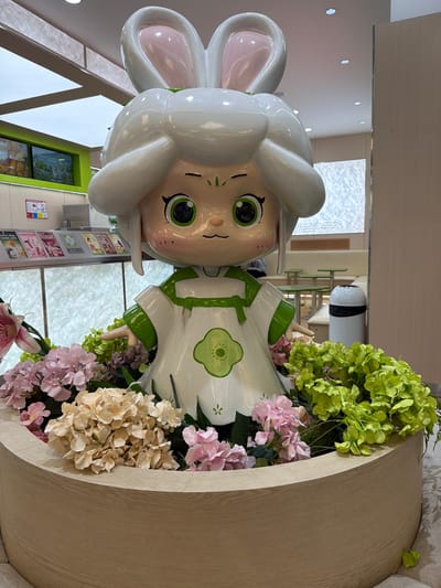 A life-sized, cartoonish statue of a character resembling a child with bunny ears and a white dress with green trim is positioned in the center of a circular wooden planter filled with various artificial flowers in shades of pink, beige, and green. The statue has large green eyes and blush marks on its cheeks. In the background, a brightly lit interior space is visible, featuring white ceiling panels with recessed lighting, green accent walls, and shelves displaying products. Further in the background, green tables and chairs suggest a seating area. The overall scene appears to be an indoor commercial space, possibly a cafe or retail store, located in Ulaanbaatar, Mongolia.
