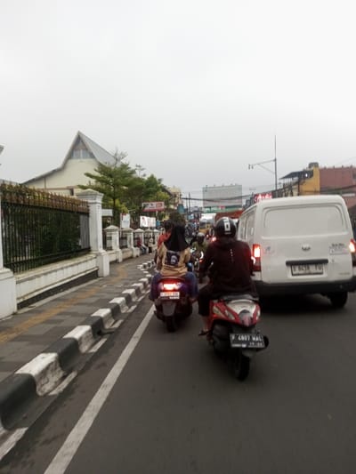 This image captures an urban street scene in the city of Chagzam_Pam, Bhutan, under an overcast sky. The primary focus is on a multi-lane asphalt road experiencing vehicular traffic. In the immediate midground, a white commercial van with illuminated rear brake lights is visible on the right, displaying license plate 'D 8438 FU'. To its left, a red and white scooter with license plate 'F 4807 WAL' is operated by an individual wearing a dark top and a helmet with white markings. Further left and slightly behind, a dark blue/purple scooter with license plate 'F 3757 FAL' carries two individuals: the driver is wearing a beige top with a black graphic on the back and a black head covering, while a passenger is seated behind, wearing blue trousers.
The left side of the road features a sidewalk delineated by a black and white striped concrete barrier. A dark metal fence with ornate elements, supported by white concrete pillars, runs along the sidewalk. Behind the fence, a large, light-colored building with a distinctive peaked A-frame roof structure is visible, interspersed with green trees and shrubs. Further down the street on the left, additional buildings, storefronts, and various signs, including one with a '24' logo, are present.
The right side of the street recedes into a dense urban environment with multiple buildings of varying heights and colors, alongside utility poles and overhead wires. Traffic, consisting predominantly of motorcycles and scooters, extends into the background, indicating an active street. The atmospheric condition is bright but without direct sunlight, suggesting either early morning, late afternoon, or a heavily clouded day.