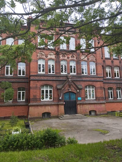 The image shows the exterior of an old, red brick building, likely a school, in Kaliningrad, Russia. The building has multiple floors with arched windows, many of which are decorated with snowflake cutouts. A large, dark double door serves as the main entrance, approached by a set of concrete stairs. Trees with green leaves partially obscure the facade. In front of the building, there is a paved area with some puddles and patches of grass, and to the left, a small fenced-off garden with bushes and a bench. The sky is overcast.