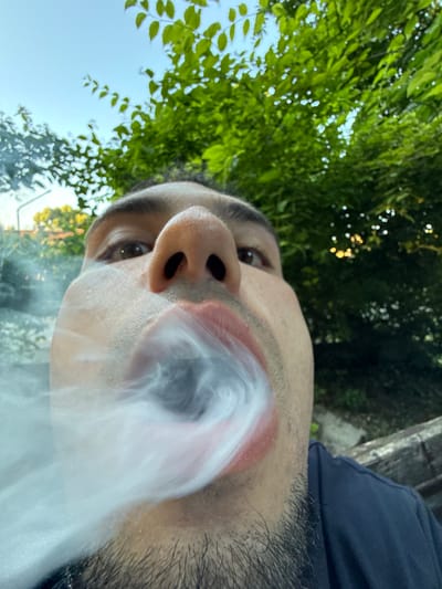 The image is a close-up, fisheye perspective selfie of a person exhaling smoke outdoors. The person's face, specifically their nose, mouth, and chin with stubble, fills the foreground. They are wearing a dark blue t-shirt. Smoke is being exhaled from their mouth, obscuring a portion of their face and creating a swirling cloud. The background consists of lush green foliage from trees and a clear blue sky. In the far background, on the left side, there are hints of buildings and trees that suggest an urban or suburban setting. The presence of a concrete bench or structure is visible on the right side of the frame. The overall lighting suggests it is daytime, possibly late afternoon or early evening due to the warm tones filtering through the leaves. The specific location in Shumen, Bulgaria is not discernable from the visual information provided in the image.