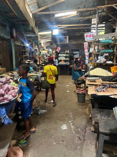 The image depicts an indoor market scene in Nkpor, Nigeria. The market appears to be a bustling place with stalls selling various goods. In the foreground, a young boy in a blue and black t-shirt and blue shorts stands looking to his left. To his right, a woman in a yellow t-shirt and red and black patterned leggings walks away from the camera, carrying a dark bag. Behind them, other people are visible, some interacting with stalls and others walking through the market.
The stalls are constructed from wood and corrugated metal, with shelves stocked with bags of grain, bottled liquids, and other packaged goods. One stall has a sign that reads "POS AVAILABLE". In another area, produce like onions is displayed in bowls and on tables. The floor of the market is dirt and appears worn, with some debris scattered around. The overall lighting suggests natural light filtering in from openings in the roof and possibly some artificial lighting. The environment has a somewhat rustic and informal feel, characteristic of many open-air markets.