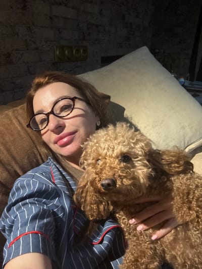 A woman with auburn hair and glasses is lying on a couch, holding a small, light brown poodle. She is wearing blue and white striped pajamas with red piping. The poodle is nestled in her arms, with its head resting on her chest. The background shows a brick wall with a decorative element, and a large, light-colored pillow. The scene appears to be indoors, possibly in a living room. There is no visible indication of the city of Dnipro, Ukraine.