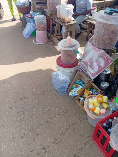 The image depicts an outdoor market scene in New Stadium, Nigeria. In the foreground, a red crate holds several bottles of dark liquid, likely beverages. To the left of the crate, a bucket contains multiple small cups filled with orange and yellow colored desserts or jellies. Behind this, a cardboard box is overflowing with various packaged snacks and candies. Further back, several large plastic containers are visible; one is filled with fried food items, another holds more of the same, and a third, smaller container also contains fried food. A person wearing bright green pants and dark sandals stands to the far left, partially obscured by a large bag. Another person, wearing a grey t-shirt and light-colored pants, is seated on a wooden stool in the background. The ground is paved with asphalt.