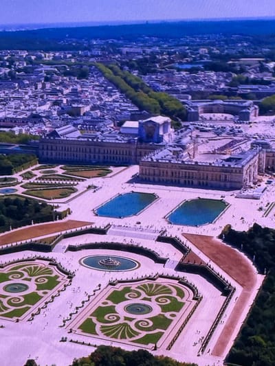 This is an aerial photograph of a vast palace complex with formal gardens. The palace is a large, ornate building with multiple wings and a grand facade. In front of the palace, there are two large rectangular pools. Beyond the pools, the gardens are meticulously designed with manicured hedges, symmetrical patterns, and fountains. The scene is bathed in daylight, and there are many small figures of people visible in the gardens, suggesting a public space. The architecture and garden design are characteristic of European royal residences. This is a view of the Palace of Versailles in France, not Dubai.