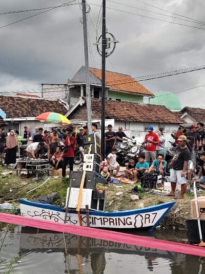 The scene depicts a group of people gathered near a canal in the Kecamatan Tanggulangin area of Indonesia.  The canal is lined with green vegetation. A decorated boat with the words "CIMPRING R7" written on it, is partially submerged in the canal. The boat is laden with what appears to be speakers or musical equipment.  The group of people consists of men, women, and children. Some are standing around, while others are seated. A woman in a hijab is standing near a blue motorbike. A man wearing a blue shirt is holding a microphone. There is a power pole in the center of the scene with power lines strung overhead. The sky is overcast with gray clouds. The scene is reminiscent of a community gathering.