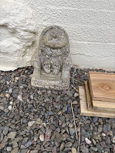 A stone Buddha statue sits adjacent to a white exterior wall and on a bed of gray gravel. Two pieces of light brown wood are placed near the gravel and the wall. The scene is outdoors, presumably in Longden, United Kingdom.