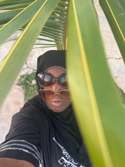 This is a vertical, close-up, eye-level shot of a Black person, viewed from above and slightly to the side, with large green palm fronds framing the top and left of the image. The person is wearing a black hijab and large, round sunglasses with brown lenses. They are looking down and slightly to the right, with their mouth open and tongue visible. The person is wearing a black garment with white decorative stitching along the neckline and sleeves. The background is blurred, showing a light-colored concrete surface with some sparse green vegetation. The lighting suggests it is daytime, possibly sunny. The overall impression is casual and intimate, perhaps a selfie taken outdoors in a tropical or subtropical environment. No text is visible in the image. The location context is Yelwa, Nigeria.