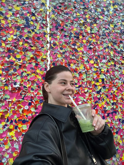 An adult female is depicted in a medium shot, facing slightly to her right, with a gentle smile while drinking from a clear plastic cup through a white straw. Her dark hair is pulled back, and she is wearing a black leather jacket. Her right hand holds the cup, which contains a green liquid, possibly a matcha beverage, approximately half full. Her fingernails are manicured with a light base and darker tips. The background is a vertical wall densely covered with numerous overlapping, colorful circular paper discs in shades of pink, yellow, blue, green, orange, and purple, creating a vibrant, textured surface. A thin, vertical reflective strip or seam is visible in the background, slightly left of center. This distinctive colorful wall is identifiable as the "Mur des souhaits" (Wall of Wishes) at La Piscine - Musée d'Art et d'Industrie André Diligent in Roubaix, France.