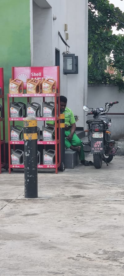 A man wearing a green shirt and green pants sits on a dark gray step to the right of a red shelf displaying Shell Helix Ultra motor oil bottles. He is looking at his phone, which he holds in his right hand. A motorcycle is parked to his right, with a license plate visible. The background includes a white building with a green wall to the left. The scene appears to be outdoors, possibly at a gas station or service center in Moba, Nigeria.