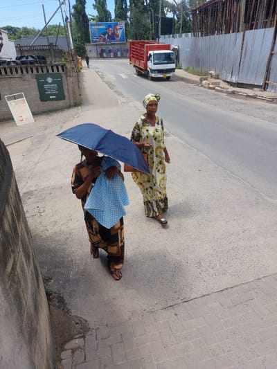 Two women are walking on a paved road. One is carrying a blue umbrella. The other wears a patterned yellow and white dress and a head wrap. A white truck with a red cargo bed is parked on the left side of the road. A building with a green sign that reads "AMBULANCE/PEDESTRIANS GATE GETI LA WAENDA KWA MIGUU/AMBULANCE" and the text "KARIBU" and "WELCOME" can be seen on the left. A billboard for Pepsi with the text "CHUPA NI MWAMBA" and "600" is also visible on the left. The scene appears to be in Dar es-Salaam, Tanzania, due to the Swahili text on the sign and billboard.