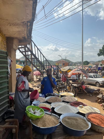 This image depicts a vibrant outdoor market scene in Gwong, Nigeria. The primary subjects are people and various food items displayed in large bowls and baskets. A person wearing a yellow head covering and a grey shirt with a floral pattern is seated at a stall, interacting with their wares. Another person in a blue and yellow t-shirt stands nearby, possibly a vendor or shopper. The market is set up along a dusty road with a backdrop of buildings and a gently sloping landscape, suggesting a hilly terrain.
Numerous large bowls are filled with different types of grains, legumes, and seeds, including what appears to be rice, beans, and peanuts. A bright green basket holds smaller plastic bags of goods. A red bucket is also visible. In the background, a silver car is parked, and several market stalls with colorful awnings and umbrellas are set up. A metal staircase leads up to an upper level of the market or an adjacent building.
The time of day appears to be daytime, with bright sunlight casting shadows and illuminating the scene. The sky is blue with scattered white clouds. The atmosphere seems busy and bustling, typical of a local market. Visible text includes "PHARMACY" on a sign in the background. The overall impression is one of lively commerce and community activity.
