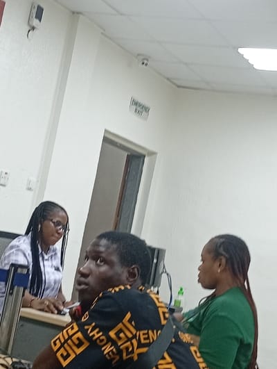 The image captures an indoor scene, likely within an office, reception area, or waiting room. Three individuals are visible, along with several objects and architectural features.
In the left foreground, a woman with dark, braided hair and glasses is seated at a desk or counter. She is wearing a light-colored plaid shirt and appears to be looking down at something on the desk, with her hands resting on the surface. Her posture suggests she is engaged in an activity at the desk.
In the center-foreground, a man with short, dark hair is seated, looking upwards and to his right, away from the camera. He is wearing a black short-sleeved shirt with a repeating yellow or orange pattern, which appears to be text, possibly "GIVENCHY". A black strap, possibly from a bag, crosses his shoulder.
To the right of the man, a third woman is partially visible from the side. She has long, reddish-brown braids and is wearing a green long-sleeved top. She is facing towards the man in the center, seemingly engaged in a conversation or observing something in that direction.
Behind the man, a computer monitor or screen is partially visible, along with some cables. A small bottle with a green cap, possibly hand sanitizer, is also on the surface behind him.
The room features light-colored (white or off-white) walls and a white drop-ceiling with a visible grid pattern. High on the wall in the upper-middle of the frame, there is a sign that reads "EMERGENCY EXIT" above a dark doorway. Two small, white surveillance cameras (CCTV) are mounted on the ceiling or high on the walls; one is visible near the top left corner, and another is visible near the top middle, above the emergency exit sign. On the far left wall, a white square device, possibly another sensor or a network access point, is mounted near the ceiling, and an electrical outlet is visible lower down.
The lighting in the room is bright and even, consistent with artificial indoor lighting. The overall atmosphere appears calm, with the individuals either interacting or waiting.