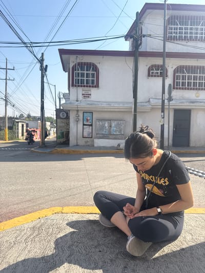 A medium-shot, slightly rotated photograph shows a woman squatting on a sidewalk next to a street in Heroica Puebla de Zaragoza, Mexico. The woman is wearing black leggings, a black t-shirt with a yellow design, and gray sneakers. She has her head bent down and her hands clasped together. The sidewalk is gray and textured, with a yellow curb separating it from the street.

Behind the woman is a two-story white building with dark red trim around the windows. The windows have metal bars. There are signs and posters on the building. Overhead, power lines run in multiple directions. In the background, the street stretches into the distance with other buildings and greenery visible. The sky is clear and blue.