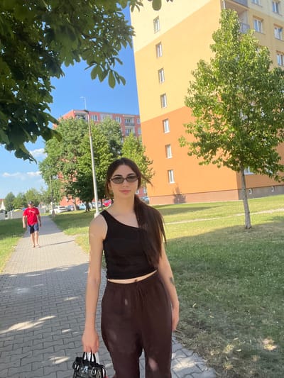 A young woman with long dark hair and sunglasses is posing for a photo outdoors. She is wearing a black sleeveless top and dark leggings. She is standing on a paved walkway next to a grassy area. In the background, there are apartment buildings and trees. The sun is shining, suggesting it is daytime. The scene appears to be in a residential area. Another person, a man in a red shirt and blue shorts, is walking in the distance.