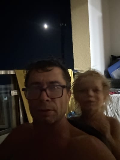The image is a selfie taken at night, likely outdoors, with a moon visible in the dark sky. In the foreground, a man wearing glasses is looking at the camera. Behind him, a child with blond hair is partially visible and out of focus. The background shows a dark blue curtain or blind, a textured wall, and some indistinct objects, possibly furniture or luggage. The man appears to be shirtless. The overall lighting suggests it is nighttime. There is no indication of the specific location within Boreti, Montenegro.