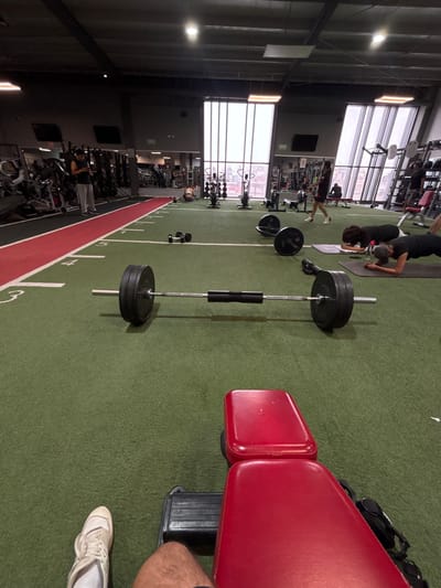 This media file captures the interior of a modern gym in Cholula, Mexico, from a slightly elevated perspective, looking across a spacious exercise area.
In the foreground, a vibrant red gym bench is prominent on the right. A person's leg and white athletic shoe are partially visible on the far right, suggesting someone is seated or standing nearby. Central to the image is a barbell, loaded with multiple black weight plates; one plate clearly shows a "25KG" marking. The floor beneath is a green turf surface, marked with white lines and numbers like "5," "4," and "3," indicating designated zones or distances for exercises.
In the mid-ground, two individuals are actively exercising on dark yoga mats on the green turf. One person, wearing a black top and cap, is performing a plank or similar bodyweight exercise. Further into the background, the gym transitions to an area with extensive workout equipment, including various weight machines, dumbbell racks, and what appear to be treadmills or cardio machines. Several other gym-goers are visible utilizing this equipment.
The setting is characterized by a high ceiling with exposed structural elements and numerous large windows spanning the upper sections of the walls. These windows allow significant natural light to illuminate the space, suggesting it is daytime. The bright lighting through the windows blurs the view of the exterior. The overall scene depicts an active, well-equipped, and spacious fitness facility. A green sign, possibly indicating an exit, is faintly visible in the background.