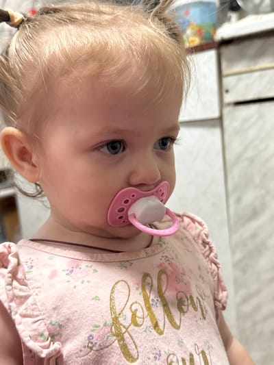 A close-up, vertically oriented shot shows a baby girl with blonde hair and blue eyes. She has a pink pacifier in her mouth. The baby is wearing a light pink shirt with a floral pattern and ruffles around the neckline and sleeves. The shirt has gold cursive text on it, which appears to read "Mommy's Little". The background is blurred, but suggests an indoor setting with light-colored walls and some shelving. The lighting is bright, possibly indicating daytime. The baby's expression is calm.