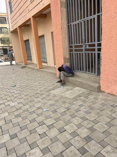 This image depicts an outdoor urban environment in Sabon Gari East, Nigeria. In the foreground-right, a male individual is seated on a concrete step against a building featuring a peach-colored stucco facade. He is dressed in a dark blue long-sleeved shirt, light grey trousers, and flip-flops, with his head bowed towards his knees. Adjacent to him is a grey metal security gate secured with a red padlock. Two light-colored wrappers are present on the step near his feet. The ground is covered with interlocking grey concrete pavers, upon which several pieces of litter, including green and white plastic wrappers, are scattered. To the mid-left, a second male individual, wearing a light grey long garment, walks away from the viewer. Behind this individual, a beige building with three prominent light orange columns and three closed storefronts with metal roll-down shutters is visible. In the background, further urban structures and partially visible vehicles indicate a street setting. The scene is under bright daylight.