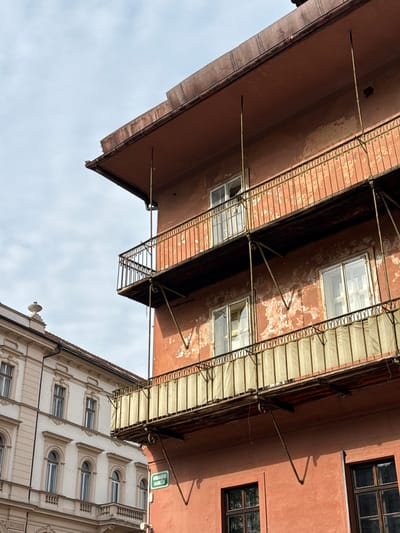Low-angle view of buildings against a cloudy sky in Ljubljana, Slovenia. Two buildings are prominent: a red-colored building with balconies extending outward, and a white ornate building with arched windows. The red building shows signs of age with peeling paint. Support bars extend from the building to secure the balconies. The white building features decorative stone elements and symmetrically arranged windows. A green street sign "RIBARJEVO NABREŽJE" is visible near the white building.