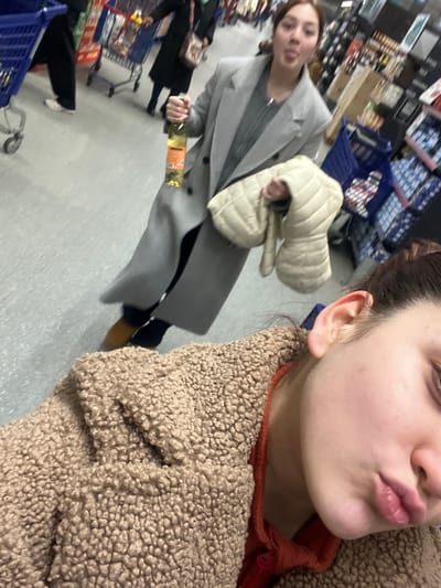 The image depicts an indoor scene within a supermarket aisle in Schiltigheim, France. In the immediate foreground, the right side of an individual's face is visible, wearing a light brown textured "teddy" coat over a reddish garment, with lips slightly pursed towards the camera, indicating a selfie perspective. In the midground, a second individual, appearing female, stands facing the camera while sticking out their tongue. This person wears a long, light grey overcoat, dark trousers, and tan-colored boots. They hold a clear glass bottle containing a light yellow liquid and an orange label in their right hand. A folded, light beige quilted jacket is held over their left arm. The background shows a supermarket environment with multiple blue shopping carts, other out-of-focus shoppers, and stocked shelves lining the aisles under fluorescent lighting. The floor is light grey.
