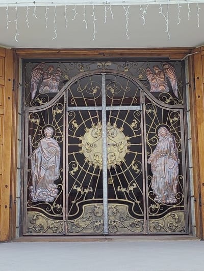 The image shows a large, ornate wrought iron gate. The gate is divided into two panels, each featuring a depiction of a religious figure. On the left panel, a male figure stands on clouds, holding a staff and a book. He appears to be a saint, possibly Saint Joseph. On the right panel, a female figure, likely the Virgin Mary, stands with her hands outstretched. Above these figures, at the top of the gate, are two angels, their faces downcast. The central part of the gate features a sunburst motif with a cross in the center. The overall design is intricate and decorative, with scrollwork and floral elements. The gate is set within a wooden frame, with wooden doors on either side. String lights are visible hanging above the gate. The scene appears to be an exterior entrance, possibly to a church or religious building, given the imagery. The lighting suggests it is daytime, with no specific weather conditions discernible. The style of the gate and the imagery suggest a European, possibly Eastern European, context. The mention of Hrodna, Belarus as a location cue further supports this. No visible text is present in the image. The subjects are static religious figures, and the activity taking place is simply the presence of the gate itself as an entrance.