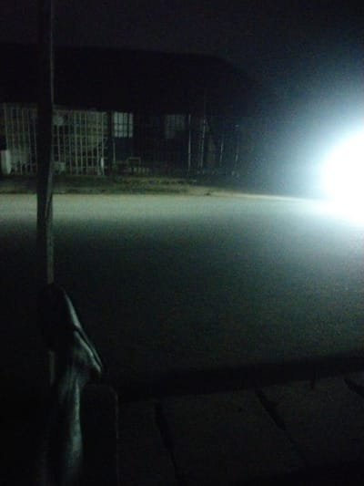 This is a low-light, slightly blurred image taken at night in Uyo, Nigeria. In the foreground, a dark, indistinct figure stands in silhouette on a wooden platform. To the left, a wooden post is visible. The midground shows a paved surface, possibly a road or parking lot, illuminated by a bright light source off-frame to the right. In the background, a building with barred windows and dark structures is dimly visible. The overall scene suggests an outdoor location, likely an industrial or commercial area, at night, with artificial lighting being the primary light source.