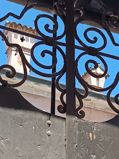 A close-up shot reveals a wrought iron gate obscuring a building in Mezzana Sant'Antonio, Italy. The gate, painted a dark color, displays ornate spiral patterns. Through the gate's openings, a light pink building with a tower is visible against a clear blue sky. The tower is adorned with a coat-of-arms or similar emblem. Bird deterrent spikes run along the building's roof. The wooden part of the gate is weathered with peeling paint.