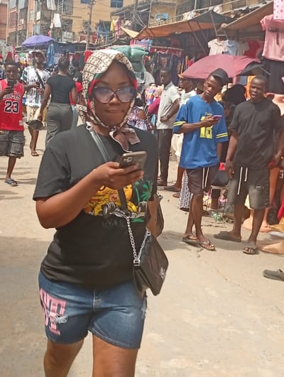 The image displays an outdoor market street scene in Fegge, Nigeria, under daylight conditions. A woman is positioned in the foreground, walking towards the viewer while looking down at a silver-colored smartphone held in both hands. She wears a cream and brown patterned headscarf with green and red accents, large dark-rimmed eyeglasses, a black short-sleeved t-shirt with a colorful graphic (featuring a yellow race car and green palm trees), and distressed blue denim shorts with pink lettering on the left leg. A black shoulder bag with a silver chain strap hangs from her left shoulder.
The background reveals a bustling urban environment. Numerous individuals, predominantly male, are present, either walking or standing. Several individuals are engaged with their mobile phones. Notable figures include a man on the left wearing a red basketball jersey with "BULLS 23," and a man in a blue t-shirt with yellow accents also looking at a phone further back. Another man in a black t-shirt stands to the right.
The street is lined with commercial activity, featuring makeshift market stalls displaying clothing and other goods under various tarpaulins and umbrellas. Multi-story buildings, some with visible balconies and clothes hanging, form the backdrop. The ground surface appears to be light-colored concrete or paved earth.