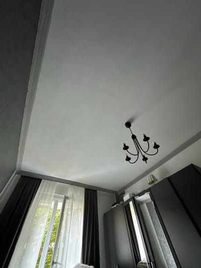 The image shows an indoor scene, likely a bedroom, looking upwards towards the ceiling. A dark, multi-armed chandelier with five candle-shaped lights hangs from the ceiling. To the left, a window is visible with dark gray curtains on the outer sides and sheer white curtains in the center, allowing natural light and greenery from outside to filter through. On the right side of the image, a portion of a large, dark wardrobe or cabinet is visible, with a small lamp with a striped shade placed on top of it. The walls appear to be painted in a light gray color, with darker gray accents around the ceiling and window frame. There are no people or animals present in the scene, and no specific actions are taking place. The image does not contain any information that would allow for its location to be determined as Hamburg, Germany.