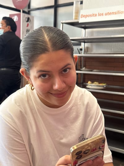A woman with dark hair pulled back and wearing gold hoop earrings is looking at the camera and smiling. She is wearing a white t-shirt. She is holding a gold-colored smartphone with a fruit-themed case in her right hand. The phone screen is not visible. A person wearing a black shirt and a face mask is partially visible in the background. The background also includes a metal structure with a sign in Spanish, "Deposita los utensilios". The scene appears to be indoors, possibly a restaurant or food establishment. The location is San Andrés Cholula, Mexico.