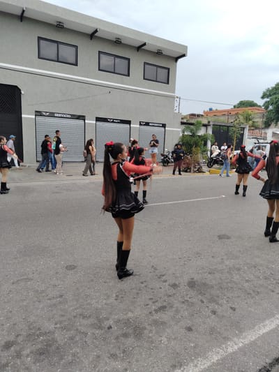 The scene is set in the city of Tinaquillo, Venezuela, on an asphalt road with buildings and people on either side. The weather is overcast. A group of young women in matching black and red outfits with boots and red bows in their hair are performing, likely a majorette or dance group. Pedestrians are on the sidewalks and streets, watching the performance. A police officer is standing on the roadside, possibly providing security. Parked motorcycles and cars are visible in the background. Buildings with gray facades and closed metal shutters are along the street. A gate and a partial view of residences are beyond the road.