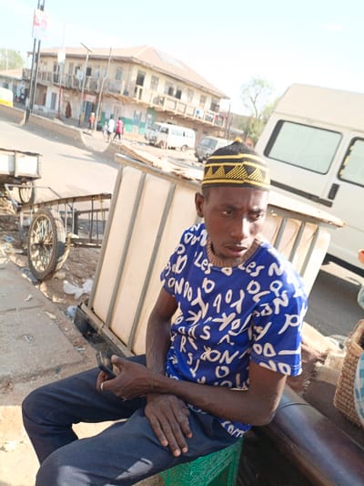 The image depicts a street scene in Gawuna, Nigeria, under bright daylight. In the foreground, an adult male with dark skin is seated on a green plastic crate. He is wearing a blue short-sleeved t-shirt with scattered white letter patterns, dark trousers, and a black kufi-style cap with yellow geometric designs. A light-colored beaded necklace is visible around his neck, and a thin green bracelet is on his left wrist. He holds a black smartphone in his right hand, with his left hand resting on his lap. His gaze is directed towards the viewer's left. Behind the man, a structure composed of light-colored panels and a metal frame is partially visible. To the man's left, a two-wheeled pushcart with a wooden-spoked wheel rests on the ground. The background features a paved street ascending gently, lined by a multi-story building with balconies and other structures. Multiple vehicles, including a white van prominent in the mid-ground and other vehicles further down the road, are present. Several indistinct individuals are visible walking in the distant background.