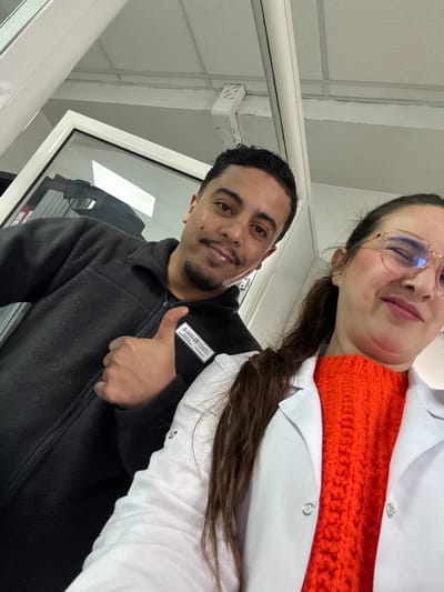 The image displays two individuals, one male and one female, posing in an indoor commercial or professional setting within Meknes, Morocco. The male, positioned to the left, has dark hair, a mustache, and a short beard. He is wearing a dark, zippered fleece jacket with a white identification badge on his right chest, which reads "A. Hajjaj & F." and additional Arabic text. He presents a faint smile and a "thumbs up" gesture with his right hand. The female, positioned to the right, has dark hair tied back and is wearing light-framed eyeglasses. She is smiling and dressed in a bright orange, cable-knit sweater underneath a white lab coat or professional jacket. The background consists of a white ceiling with exposed conduits and a white-framed door or window to the left. The environment is brightly illuminated by artificial lighting.