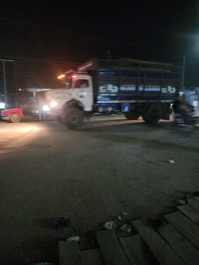 A blurry image depicts a vehicle with a trailer on a road, possibly at night in Obiaruku, Nigeria. The trailer has signage in an unknown language. The environment is dark and indistinct, with limited visibility. The image appears to be taken from a low angle.